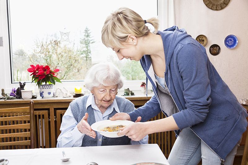 A 20 to 30 year old white female is assisting a seated senior woman with eating.
